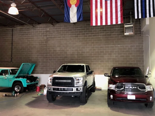 Repair bay with a classic truck, an F-150 and a Ram under Colorado and US flags